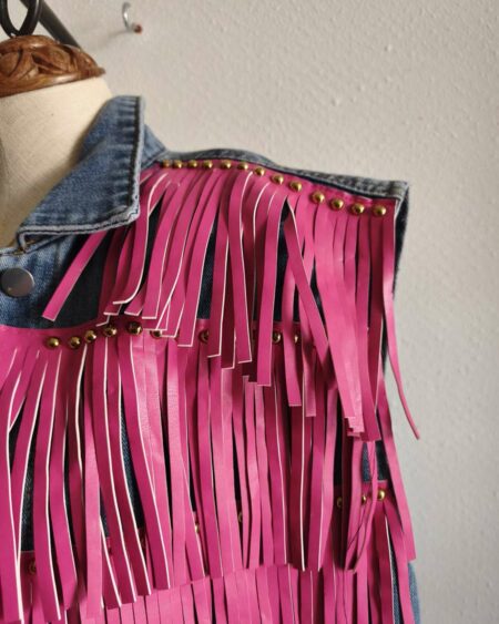 Originale veste sans manches en jean LM Lulu revisitée avec de larges franges roses et clous dorés qui lui donnent un style audacieux et très mode. Sa coupe ample permet de la porter facilement sur une robe, un top ou une chemise pour apporter une touche rock et bohème à une tenue. Une pièce forte qui attire immédiatement le regard.