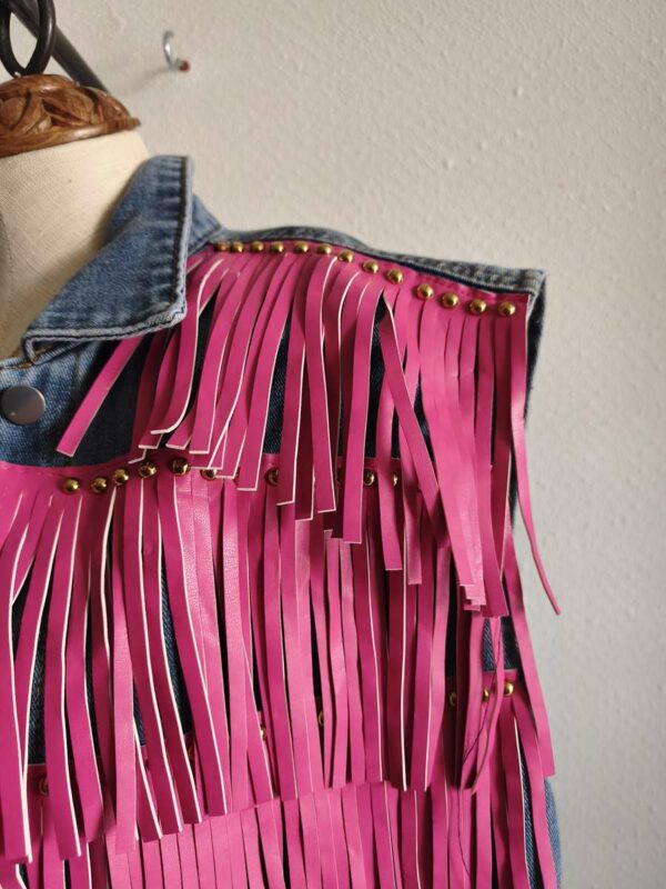 Originale veste sans manches en jean LM Lulu revisitée avec de larges franges roses et clous dorés qui lui donnent un style audacieux et très mode. Sa coupe ample permet de la porter facilement sur une robe, un top ou une chemise pour apporter une touche rock et bohème à une tenue. Une pièce forte qui attire immédiatement le regard.